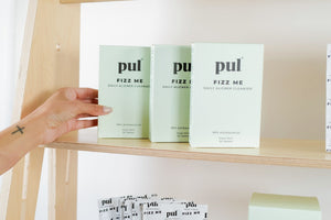 A hand reaching for a box of Pul Fizz Me daily aligner cleanser tablets neatly arranged on a wooden shelf.